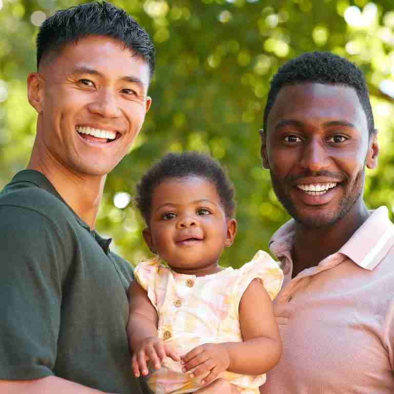 Portrait Of Same Sex Male Couple Or Friends Cuddling Baby Outdoors In Garden Or Countryside